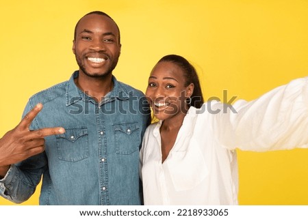 Cheerful African American Spouses Posing Making Selfie Together Gesturing V-Sign And Smiling To Camera Standing Having Fun In Studio Over Yellow Background.
