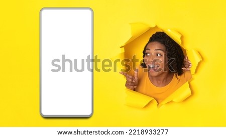 Excited African American Woman Pointing At Cellphone With Empty Screen Posing In Hole In Torn Paper On Yellow Studio Background. Great Mobile Offer Concept. Panorama, Mockup