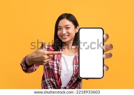 Awesome mobile app. Positive asian lady pointing at mobile phone with blank screen, advertising website or offering space for ad on yellow studio background, mockup