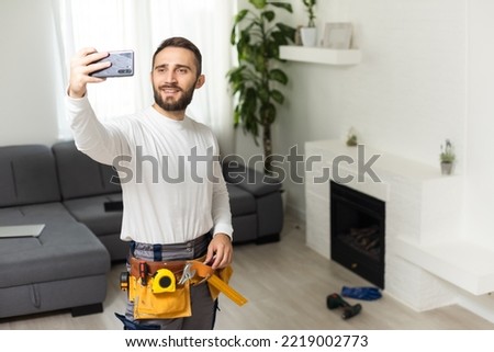 Portrait of handsome cheerful repairer shooting selfie on smart phone, wearing shirt and overalls