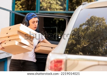 Courier carrying pizza to office by car, young woman holding boxes pile. Pizzeria delivery service employee in headphones carrying lunch, person taking out fast food from vehicle