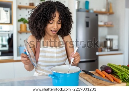 Photo of pretty African American woman holding cooking ladle spoon while cooking soup with fresh vegetables in kitchen at home