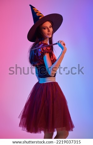 Halloween celebration. Charming young witch. Portrait of a cute teenage girl posing at studio in a witch's costume and a magic wand in her hand. Mixed colorful lighting.