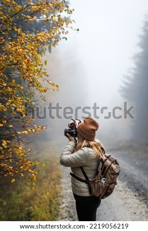 Woman with camera taking picture of autumn leaf. Tourist hiking in misty forest. Landscape photographer with backpack enjoying nature in fall season