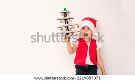 Portrait of smiling child in Santa Claus hat with handmade Christmas tree isolated on white background. 5 year happy European boy. New Year holiday creative decor idea. Cardboard craft and pasta toys.