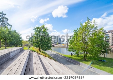 Wooden steps and seats invite you to rest in Baakenpark in Hamburg. The blue sky is streaked with clouds, the trees and meadows shine fresh green. Element of the Hafencity.