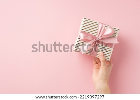 Christmas Eve celebration concept. First person top view photo of woman's hand holding stylish present box with pink ribbon bow on isolated light pink background with copyspace