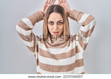 Young blonde woman wearing turtleneck sweater over isolated background doing bunny ears gesture with hands palms looking cynical and skeptical. easter rabbit concept. 