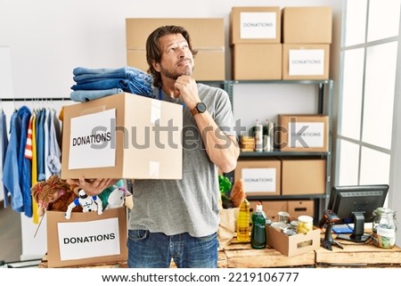 Handsome middle age man holding donations box for charity at volunteer stand with hand on chin thinking about question, pensive expression. smiling with thoughtful face. doubt concept. 