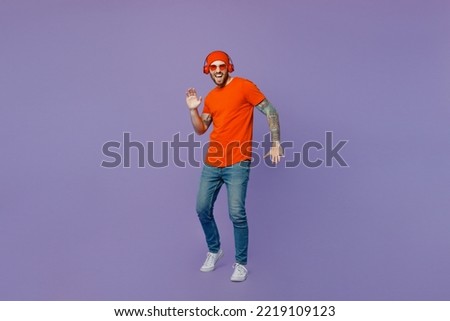 Full body young happy cool european man 20s he wearing red hat t-shirt sunglasses headphones listening music dance on party raise up hands look camera isolated on plain pastel light purple background