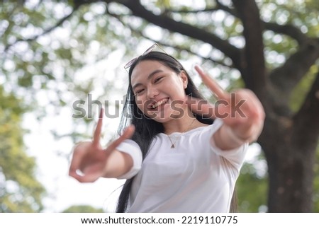 A picture of a smiling young woman holding up a peace sign with greenery in the background.