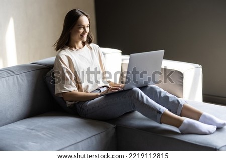 Photo of cheerful nice woman smiling and using laptop while sitting on couch in bright room