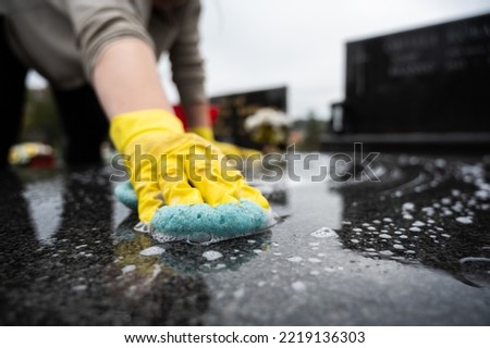Headstone cleaning on cemetery. Professional in yellow gloves cleans the marble grave, scrubbing with sponge and water. Tombstone preparation for All Saints Day on November 1st Royalty-Free Stock Photo #2219136303