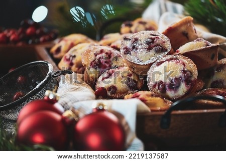 Cranberry muffins dusted with powder sugar in tray with Christmas tree and fresh berries in the background. Ornaments and vintage duster nearby. Selective focus with blurred foreground and background.