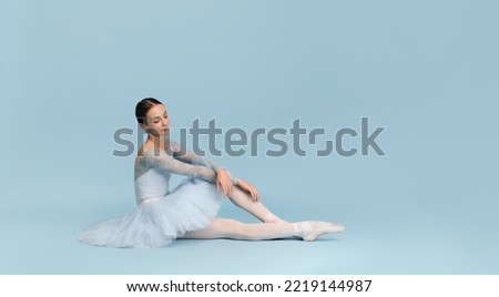 Portrait of tender young ballerina dancing, performing, sitting on floor isolated over blue studio background. Flyer image. Grace. Concept of classic ballet, inspiration, beauty, dance, creativity