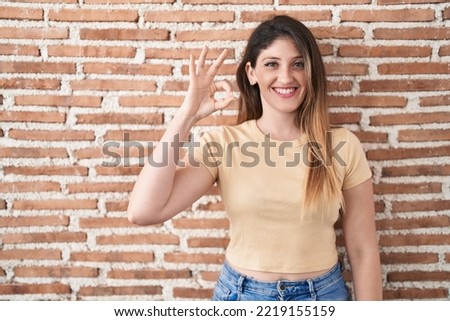 Young brunette woman standing over bricks wall smiling positive doing ok sign with hand and fingers. successful expression. 