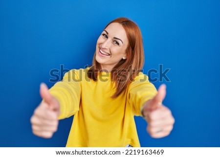 Young woman standing over blue background approving doing positive gesture with hand, thumbs up smiling and happy for success. winner gesture. 