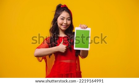 pleased asian woman holding digital tablet with green screen and showing like isolated on yellow 