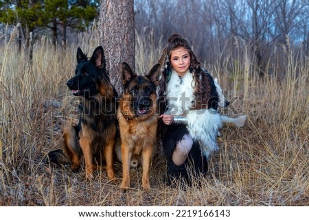 Young girl in the image of an Amazon against the background of a tree in the grass in autumn in the campaign of a pack of dogs German Shepherd and White Swiss Shepherd in the evening in the park