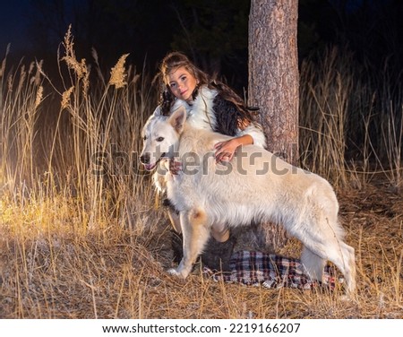 Young girl in the image of an Amazon against the background of a tree in the grass in autumn in the campaign of a pack of dogs German Shepherd and White Swiss Shepherd in the evening in the park