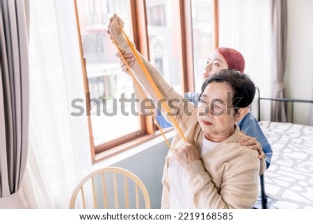 Nurse caregiver wearing scrubs exercises with a senior Asian woman by using resistance band exercise for the senior patient in physiotherapy treatment. Home health care and nursing home concept.