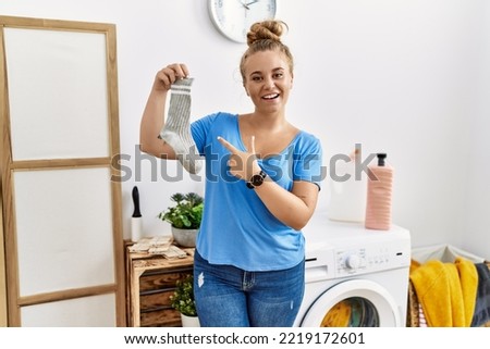 Young caucasian woman holding dirty sock at laundry room cheerful with a smile on face pointing with hand and finger up to the side with happy and natural expression 