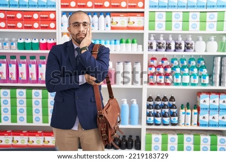 Hispanic man with beard working as salesman at pharmacy drugstore thinking looking tired and bored with depression problems with crossed arms.  Royalty-Free Stock Photo #2219183729