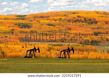Oil pump on the agricultural field in bright yellow and orange autumn. Colourful trees and blue cloudy sky are on the background.