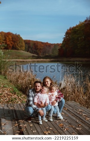 a family is walking in the middle of autumn nature with two little twin sisters; 
parents hold children in their arms and stand on a pier near a lake on a warm autumn day