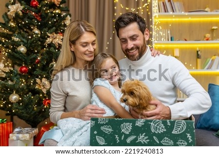 Happy parents make a surprise gift of a small dog puppy from Santa to a child for Christmas or New Year. Happiness. Father and mother surprising their kid little girl receives a present at home.