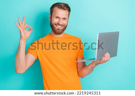 Photo of positive good mood handsome man blond hairstyle dressed orange t-shirt showing okey hold laptop isolated on teal color background