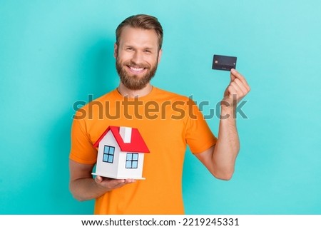 Photo of funny beard guy hold house card wear orange t-shirt isolated on teal color backgroiund