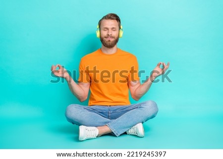 Full size photo of calm man muscular beard wear orange t-shirt jeans headphones sit in meditation pose isolated on teal color background