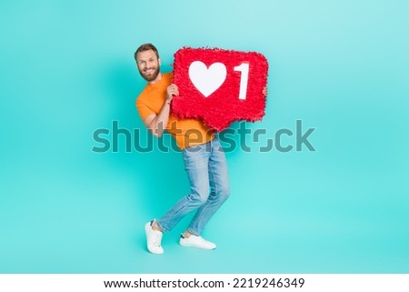 Full length photo of optimistic appreciative guy brown beard wear orange t-shirt hold big red like icon isolated on teal color background