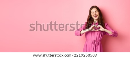 Beautiful romantic girl smiling, showing heart gesture, standing in lovely dress against pink background.