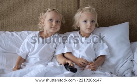Little twin sisters sit in bed leaning on pillows. They are watching TV which hangs on the wall. The screen shows children's content. From time to time they change channels.