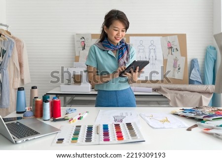 Young asian dressmaker in striped scarf checking email order from tablet computer. Laptop computer, pattern for cloth, silk thread color sample catalog and various sewing related items on the table.
