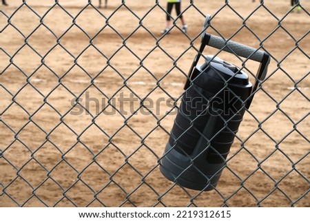 Black and gray large water jug hangs on a chain link fence. Stay hydrated during team sports.