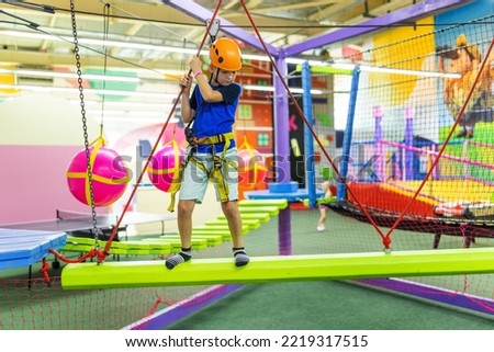 Boy in protective gear holding safety rope and passing obstacle course in indoor adventure park Royalty-Free Stock Photo #2219317515