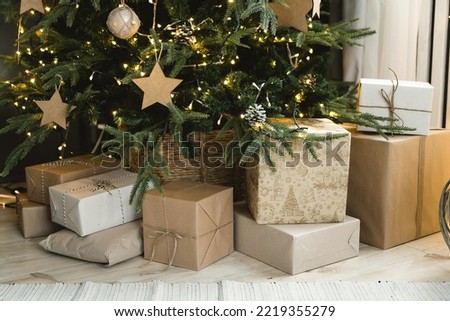 Many boxes of Christmas gifts in eco-friendly craft paper packaging and twine on the floor under a green Christmas tree decorated with garlands and craft toys. Giving gifts on Christmas morning