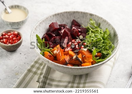 A grey deep bowl with warm winter autumn salad with arugula, red beetroot, baked pumpkin and hummus dressing cubes on a grey background