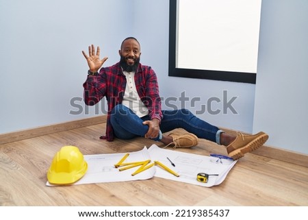 African american man sitting on the floor at new home looking at blueprints showing and pointing up with fingers number five while smiling confident and happy. 