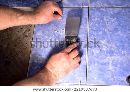 The picture shows the hands of a worker who holds a spatula in one hand. The builder is repairing the ceramic floor.