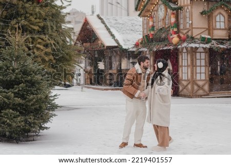 Cheerful loving couple in warm cozy clothes walk outdoors on festive city streets. Man and woman skating on ice rink as snow falls. Cold happy winter day. Holidays, Christmas, New Year, love concept.