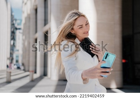 Gorgeous blonde woman in white suit walking by street smiling making selfie, talking by video call at summer day, going to work. Cheerful Italian successful businesswoman with flattering loose hair.