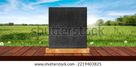 Close-up of an empty wooden table top for products display with empty blackboard against a blurred countryside landscape with green grass and daisy flowers (meadow).