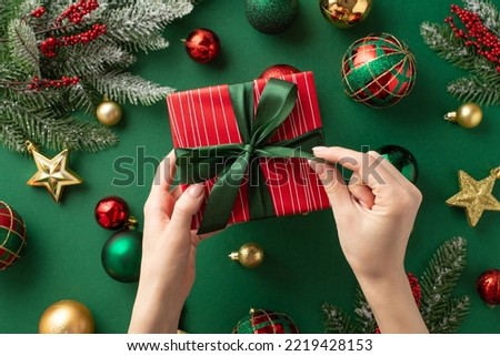 Christmas concept. First person top view photo of woman's hands untying ribbon bow on red giftbox over gold green baubles star ornaments mistletoe pine branches in snow on isolated green background