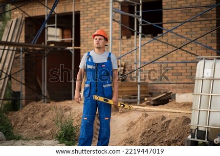 a man in a construction helmet and blue overalls holds a level in his hands and looks into the distance. High quality photo