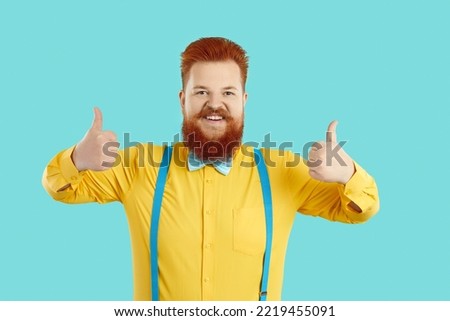 Funny, positive and stylish fat man showing two thumbs up recommending or approving your choice. Red-haired Caucasian chubby man in shirt with suspenders shows okay gesture on light blue background.