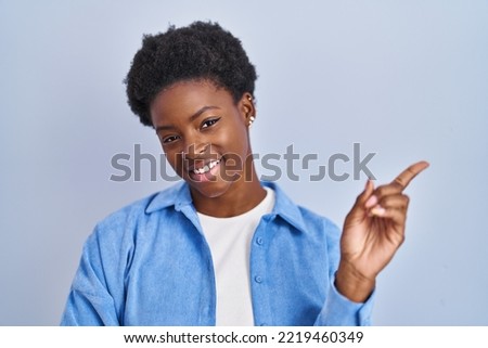 African american woman standing over blue background with a big smile on face, pointing with hand finger to the side looking at the camera. 